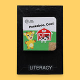 The Literacy symbol has mini children's board book titled "Let's Make Lunch" with a cartoon red bird holding corn. Background shows a picnic setting. Labeled "Literacy" in black font on white backing at the bottom of symbol . The background of the symbol is white.