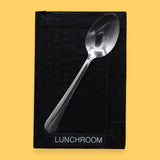 The Lunchroom symbol has a metal spoon embedded in the center. Labeled "Lunchroom" in black font on white backing at the bottom of symbol. The background of the symbol is white.
