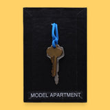 The Model Apartment high contrast tactile symbol with two keys bronze and silver on a blue ring embedded in the center. Labeled "Model Apartment" in white font on clear backing at the bottom of the symbol. The background of the symbol is black. 
