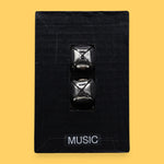 The Music symbol has two silver jingle bells attached to black felt material embedded in the center. Labeled "Music" in black font at the bottom of symbol on a white backing. The background of the symbol is white.