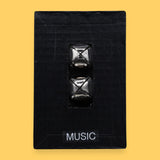 The Music symbol has two silver jingle bells attached to black felt material embedded in the center. Labeled "Music" in black font at the bottom of symbol on a white backing. The background of the symbol is white.