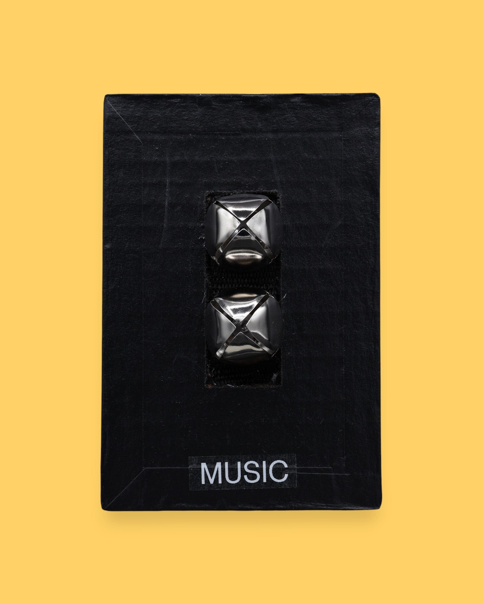 The Music symbol has two silver jingle bells attached to black felt material embedded in the center. Labeled "Music" in black font at the bottom of symbol on a white backing. The background of the symbol is white.