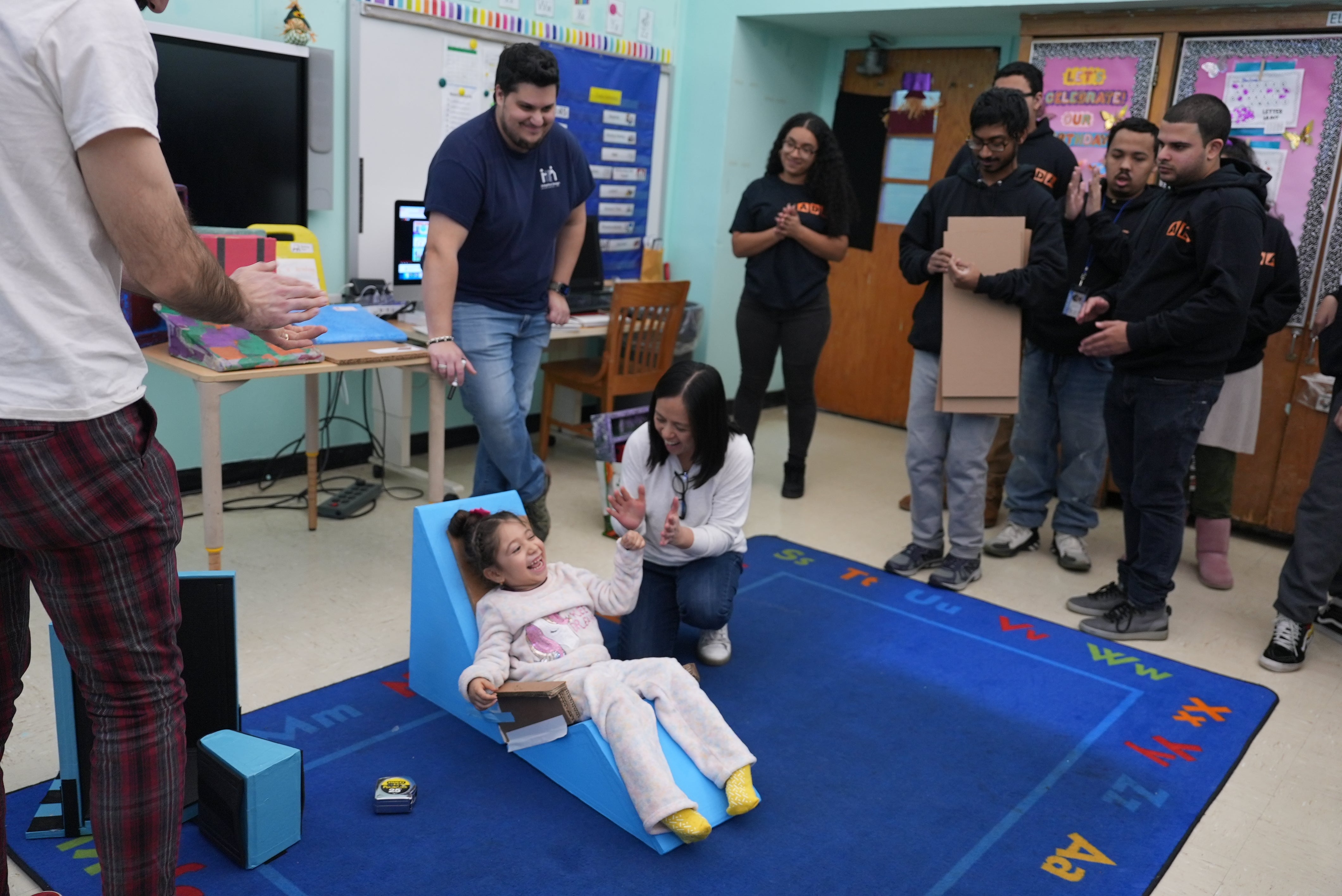 A classroom with a young child sitting on the carpeted floor. The child is leaning back in a small blue floor sitter, smiling while her Physical Therapist kneels beside her, gently supporting and encouraging her. The child is wearing light-colored clothes and bright yellow socks. Several high school student stand around, watching closely.  Their attention is focused on the child, because this is a equipment fitting for something they worked on. The Adaptive Designers are in the frame as well.