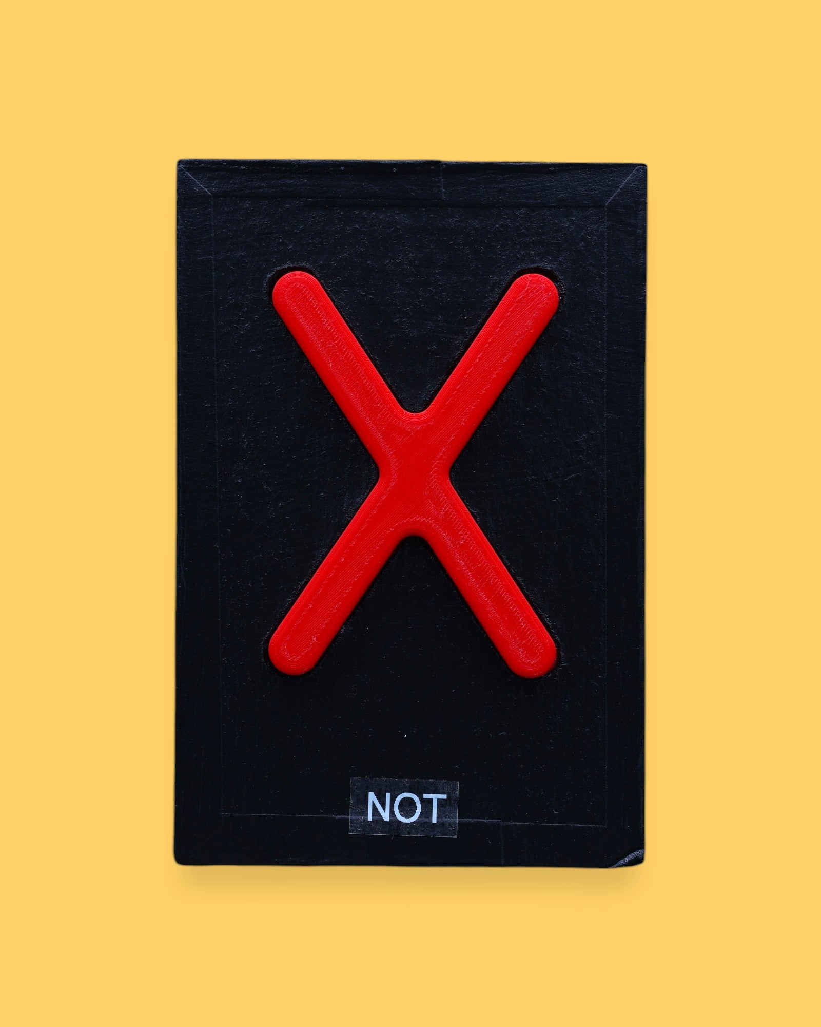 The Not high contrast tactile symbol with a bright red 3D printed filament X  embedded in the center. Labeled "Not" in white font on clear backing. The background of the symbol is black. 