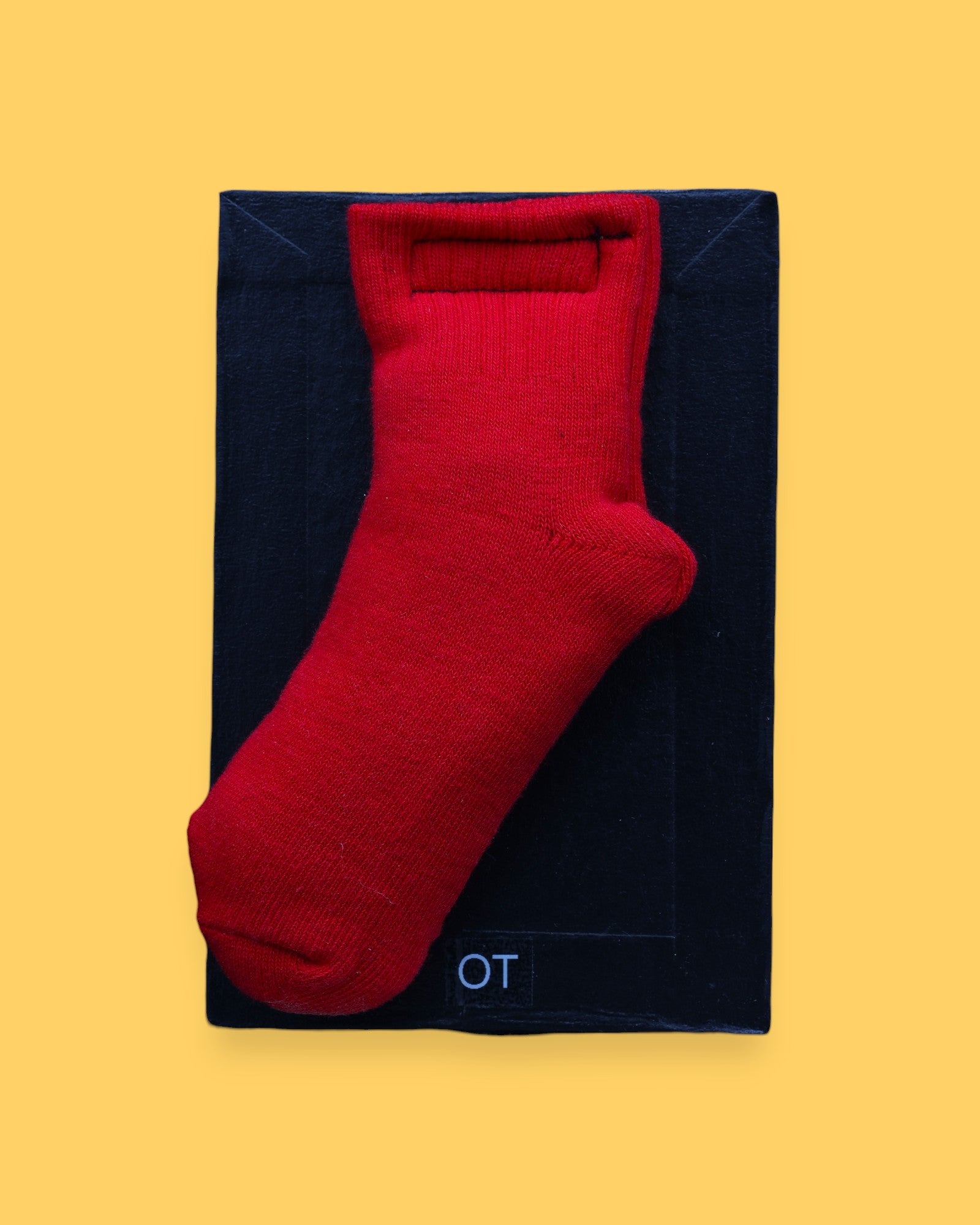 The OT Sock high contrast tactile symbol has a red cotton toddler sized sock embedded in the center with batton filling. Labeled "OT" in white font on clear backing at the bottom of the symbol. The background of the symbol is black.  