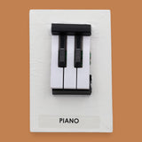 The Piano symbol with black and white hard plastic keys are embedded in a verticle position in the center of the card. Labeled "Piano" in black font at the bottom of symbol. The background of the symbol is white.