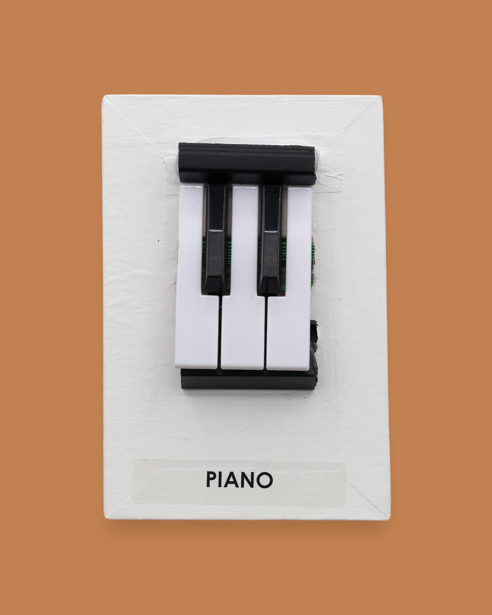 The Piano symbol with black and white hard plastic keys are embedded in a verticle position in the center of the card. Labeled "Piano" in black font at the bottom of symbol. The background of the symbol is white.
