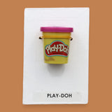 The Playdough symbol with a small yellow Play-Doh container embedded in the center. Labeled "Playdough" in black font at the bottom of symbol on white backing. The background of the symbol is white.