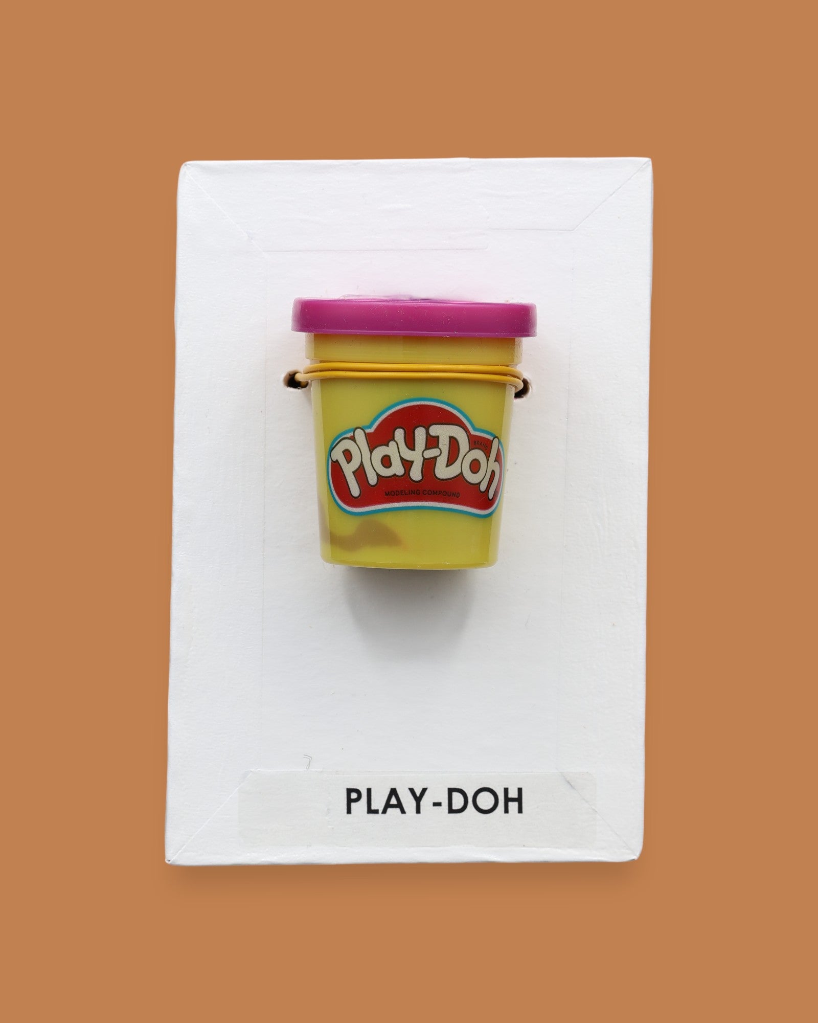 The Playdough symbol with a small yellow Play-Doh container embedded in the center. Labeled "Playdough" in black font at the bottom of symbol on white backing. The background of the symbol is white.