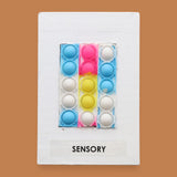 The Sensory Pop Fidget symbol with a multicolored pop-it style silicone fidget strip featuring the colors blue, pink, yellow, and white raised bubbles. Labeled "Sensory" in black font on white backing at the bottom of the symbol. The background of the symbol is white. 
