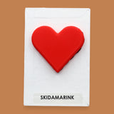 The Skidamarink symbol with a rose-red 3D printed-filament heart embedded in the center. Labeled "Skidamarink" in black font on white backing at the bottom the symbol. The background of the symbol is white. 