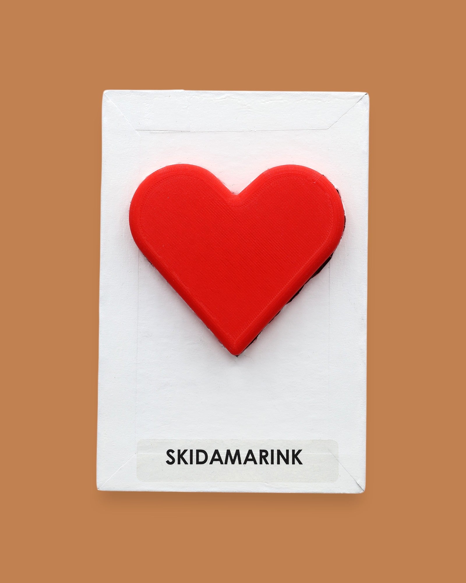 The Skidamarink symbol with a rose-red 3D printed-filament heart embedded in the center. Labeled "Skidamarink" in black font on white backing at the bottom the symbol. The background of the symbol is white. 
