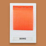 The Skiing symbol has a squared flourescent orange mesh fabric embedded in the center. Labeled "Skiing" in black font on white backing. The background of the symbol is white. 