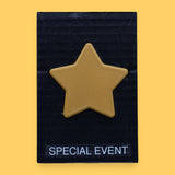 The Special Event high contrast symbol with a golden 3D printed PLA filament star embedded in the center. Labeled "Special Event" in white font at the bottom of symbol on clear backing. The background of the symbol is black. 