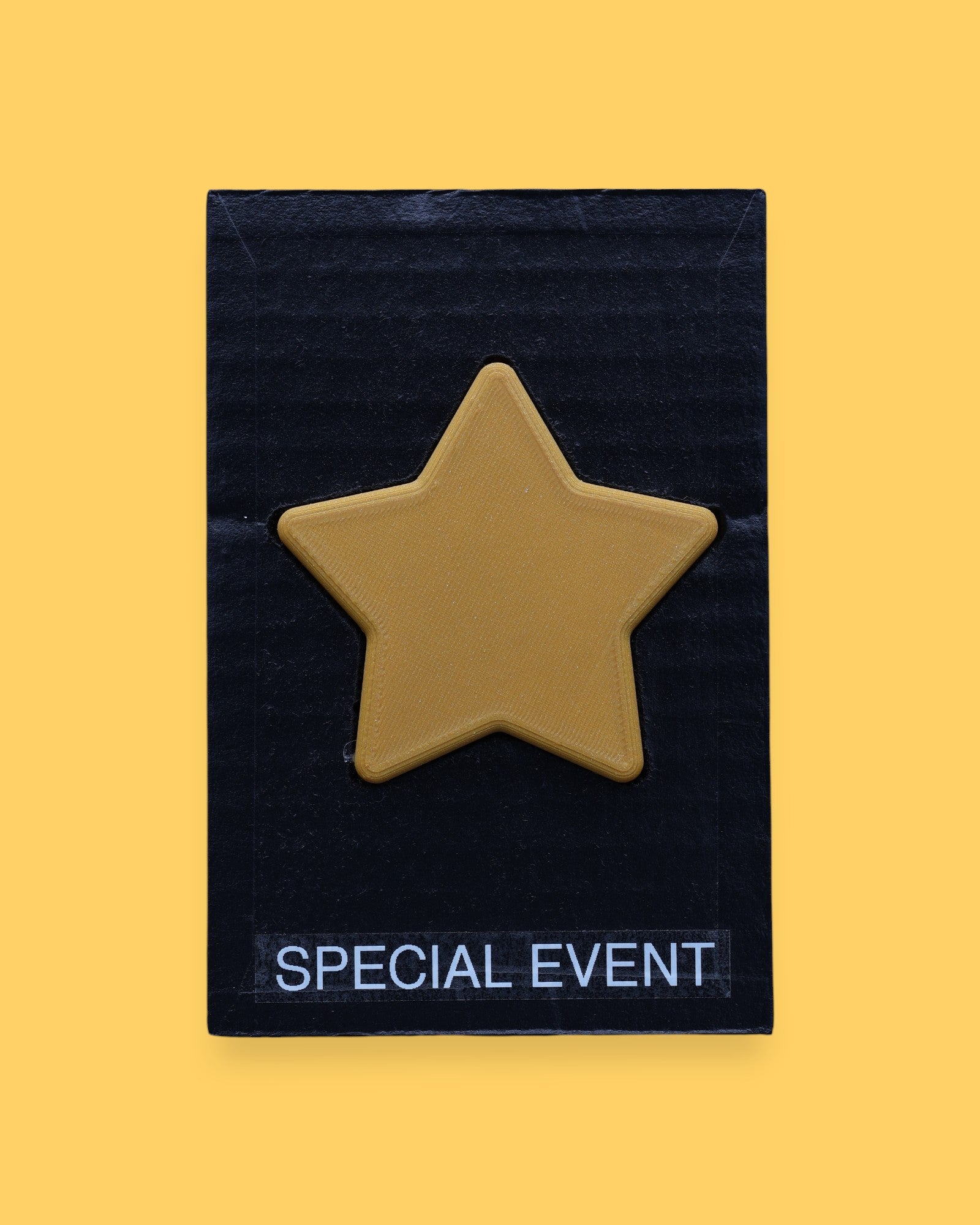 The Special Event high contrast symbol with a golden 3D printed PLA filament star embedded in the center. Labeled "Special Event" in white font at the bottom of symbol on clear backing. The background of the symbol is black. 