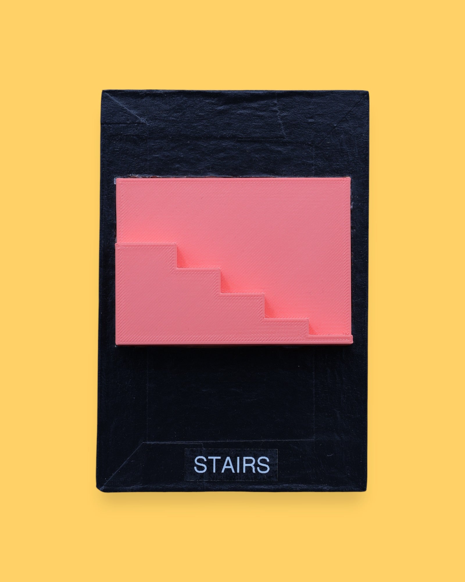 The Stairs symbol has a side profile of 3D printed stairs in bright coral orange PLA 3D printer filament. Labeled "Stairs" with black font on white backing at the bottom of symbol. The background of the symbol is white. 