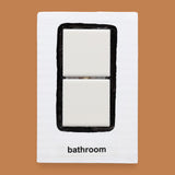 The Bathroom symbol has two square, white, ceramic tiles are embedded vertically in the center of the card with a black outline painted around the tiles. Labeled "Bathroom" in black font on white backing at the bottom the symobl. The background of the symbol is white.