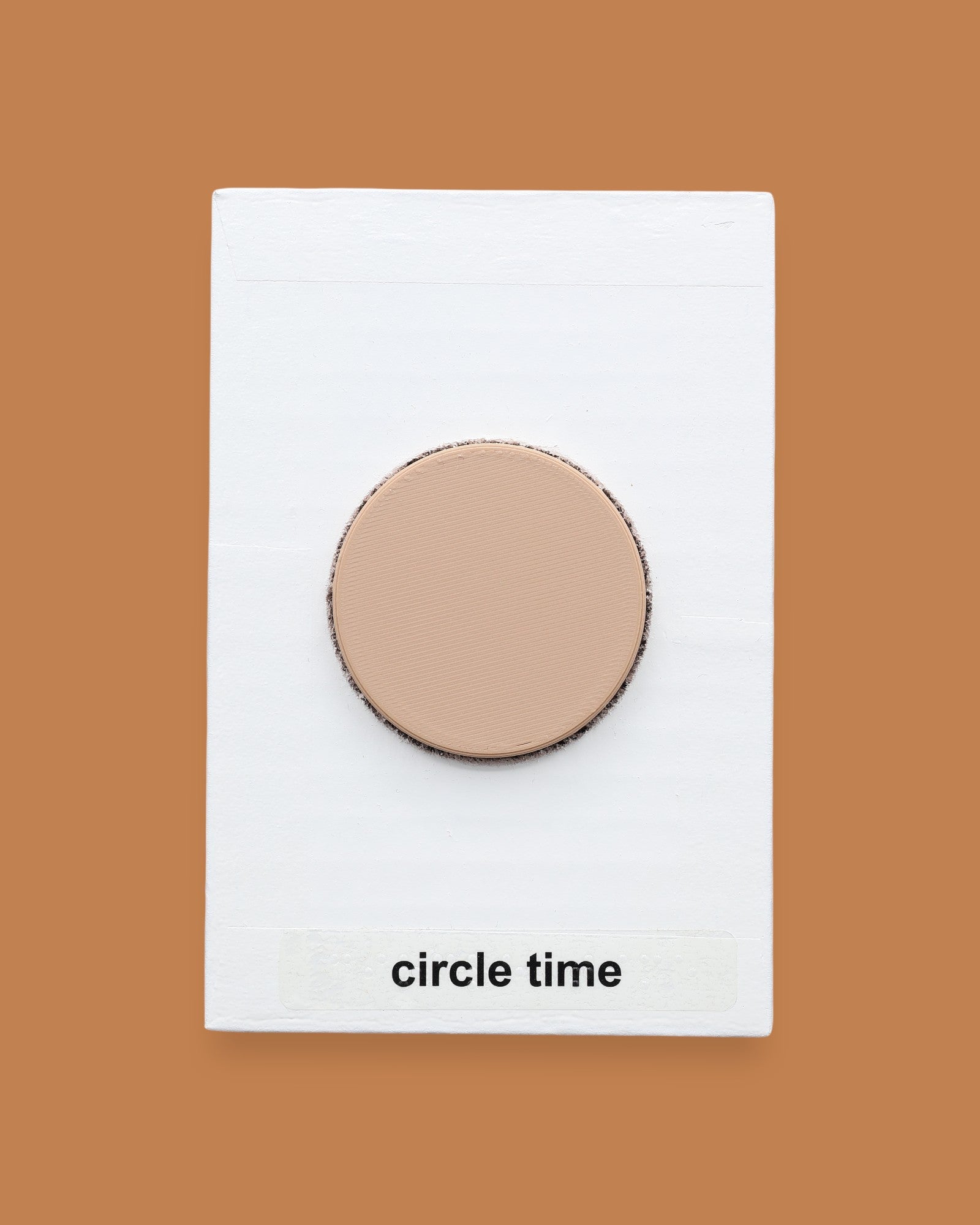 The Circle Time symbol with a tan wood-like 3D printed plastic circle embedded in the center. Labeled "Circle Time" in black font at the bottom of symbol on white backing. 