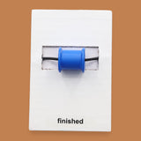 The Finished symbol has an empty thread spool, embedded horizontally in the center. The color of the spool may vary. The background of the symbol is white. Labeled "Finished" in black font at the bottom of symbol on white backing. 