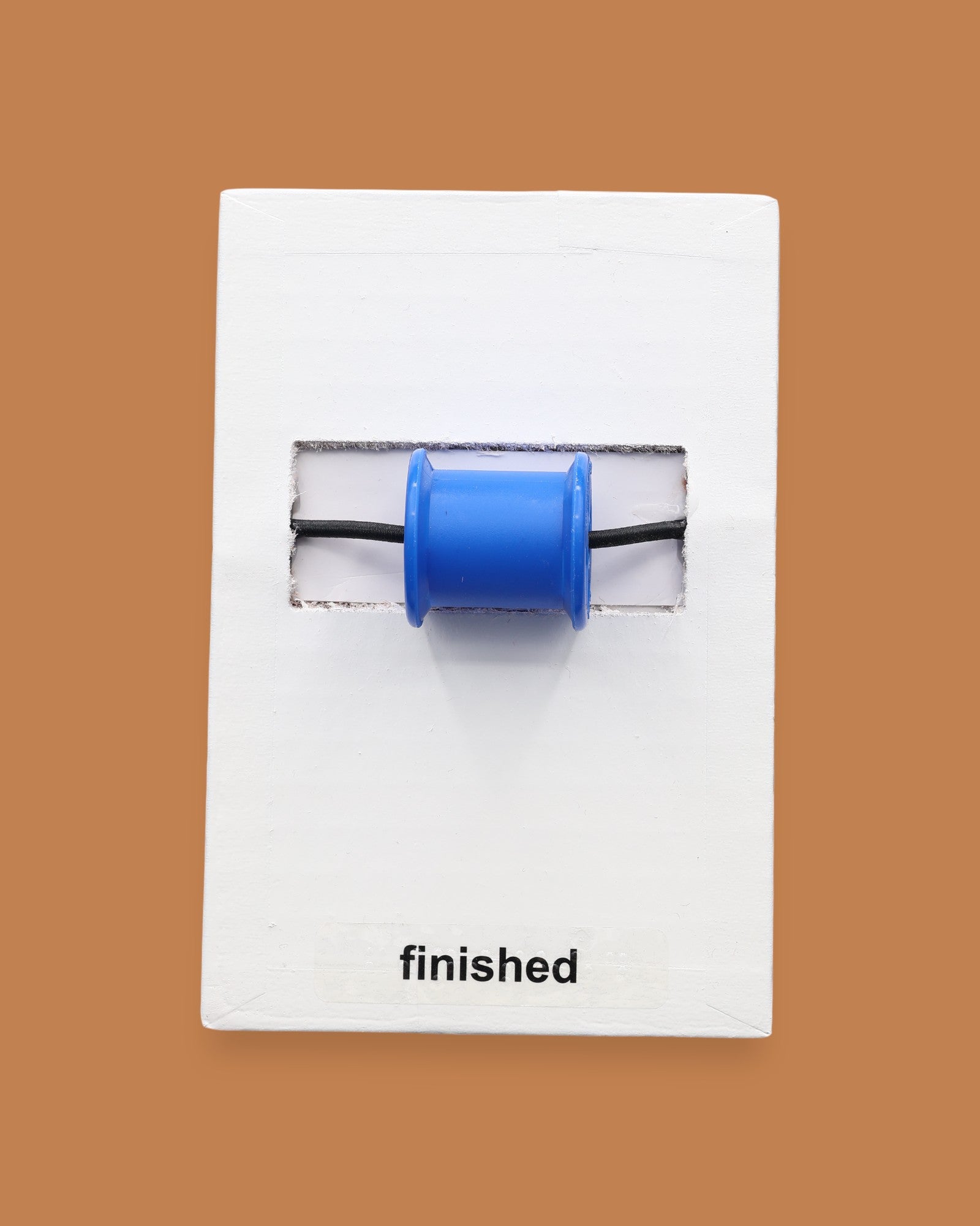 The Finished symbol has an empty thread spool, embedded horizontally in the center. The color of the spool may vary. The background of the symbol is white. Labeled "Finished" in black font at the bottom of symbol on white backing. 