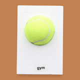 The Gym symbol has a bright yellow tennis ball is centered on a white rectangular surface against a white background. Labeled "Gym" in black font on white backing at the bottom of symbol. The background of the symbol is white.