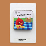 The Literacy symbol has mini children's board book titled "Let's Make Lunch" with a cartoon red bird holding corn. Background shows a picnic setting. Labeled "Literacy" in black font on white backing at the bottom of symbol . The background of the symbol is white.