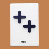 The More symbol with raised 3D PLA  navy blue PLA plus-sign shapes attached diagonally. Labeled "More" in black font at the bottom of symbol on white backing. The background of the symbol is white.