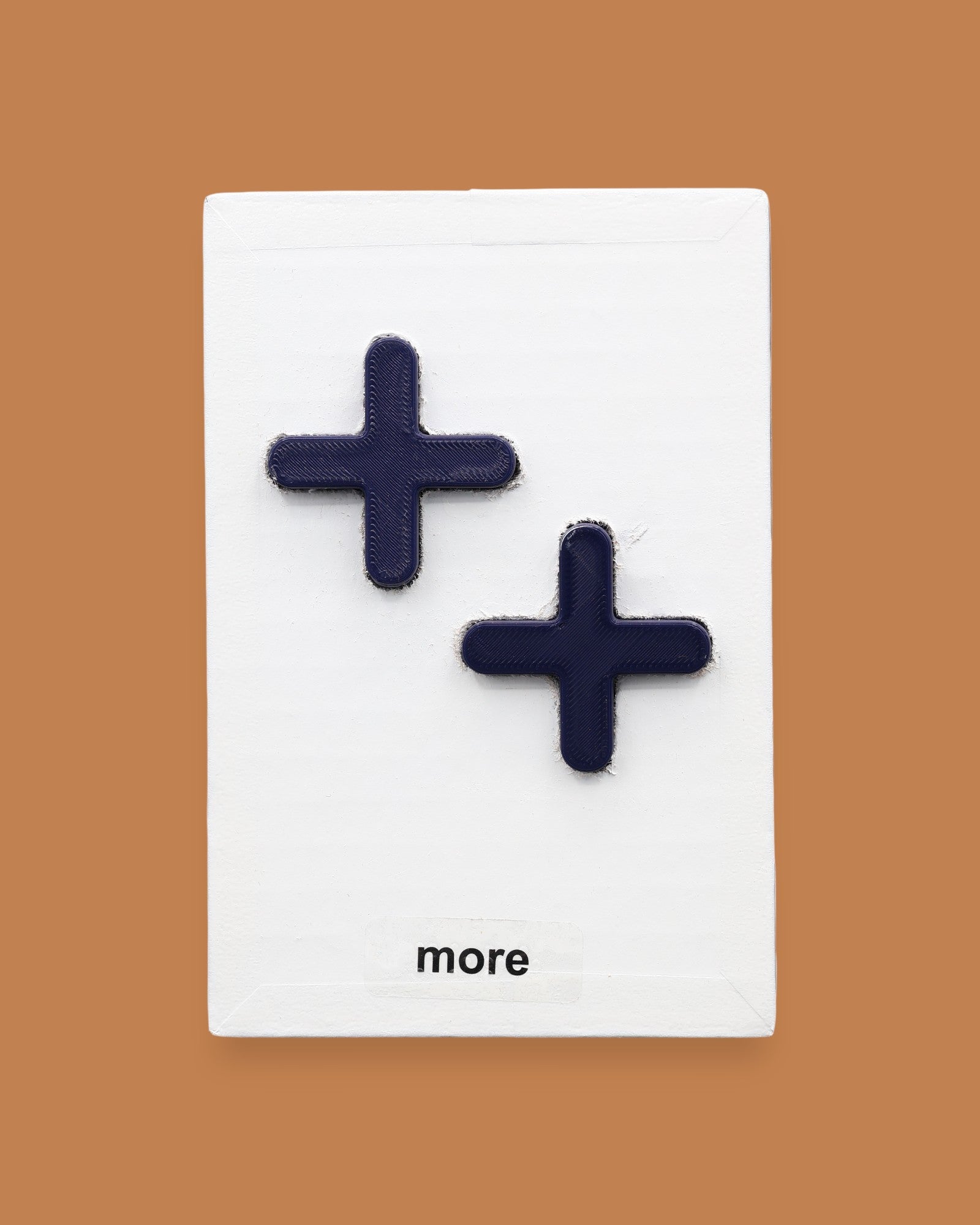 The More symbol with raised 3D PLA  navy blue PLA plus-sign shapes attached diagonally. Labeled "More" in black font at the bottom of symbol on white backing. The background of the symbol is white.