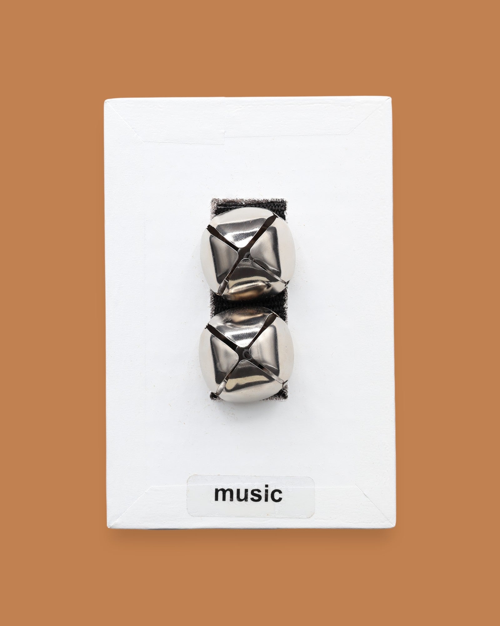 The Music symbol has two silver jingle bells attached to black felt material embedded in the center. Labeled "Music" in black font at the bottom of symbol on a white backing. The background of the symbol is white.