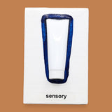 The Sensory symbol has a small white travel lotion bottle is embedded vertically in the center with a navy blue hand painted outline around the tube. The symbol has a white background. Labeled "Sensory " in black font at the bottom of symbol on white backing. 