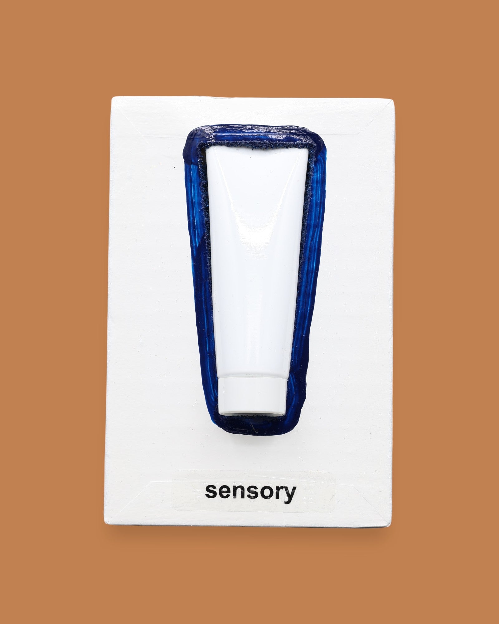 The Sensory symbol has a small white travel lotion bottle is embedded vertically in the center with a navy blue hand painted outline around the tube. The symbol has a white background. Labeled "Sensory " in black font at the bottom of symbol on white backing. 