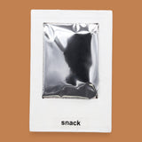 The Snack symbol with a silver mylar rectangular snack-shaped bag embedded in the center. Labeled "Snack" in black font at the bottom of symbol on white backing. The background of the symbol is white.
