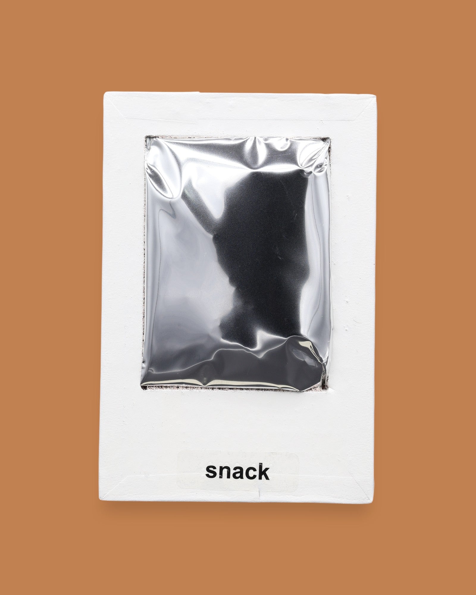 The Snack symbol with a silver mylar rectangular snack-shaped bag embedded in the center. Labeled "Snack" in black font at the bottom of symbol on white backing. The background of the symbol is white.