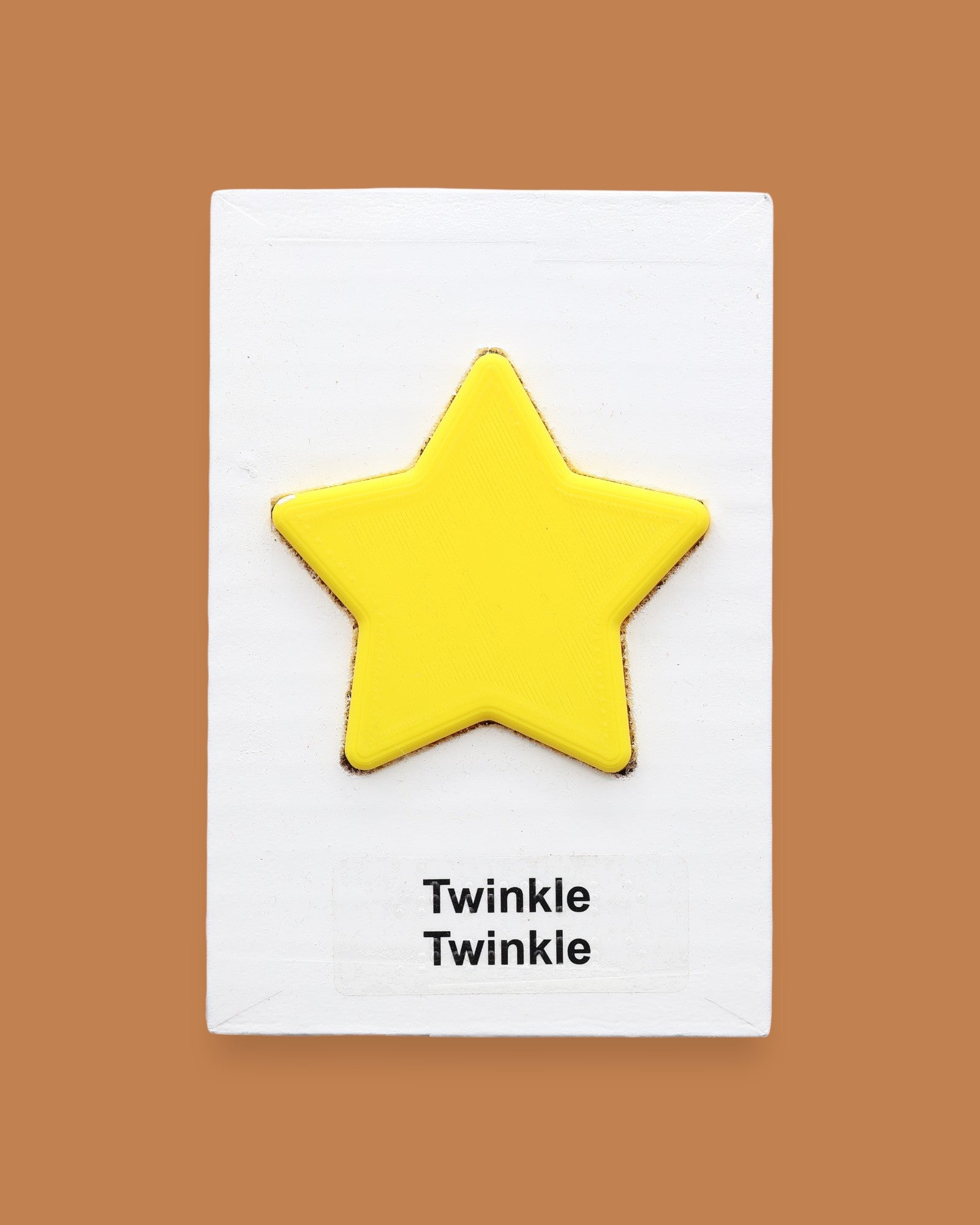 The Twinkle, Twinkle symbol with yellow 3D-printer filament star embedded in the center. Labeled "Twinkle, Twinkle" in black font at the bottom of symbol on white backing. The background of the symbol is white.