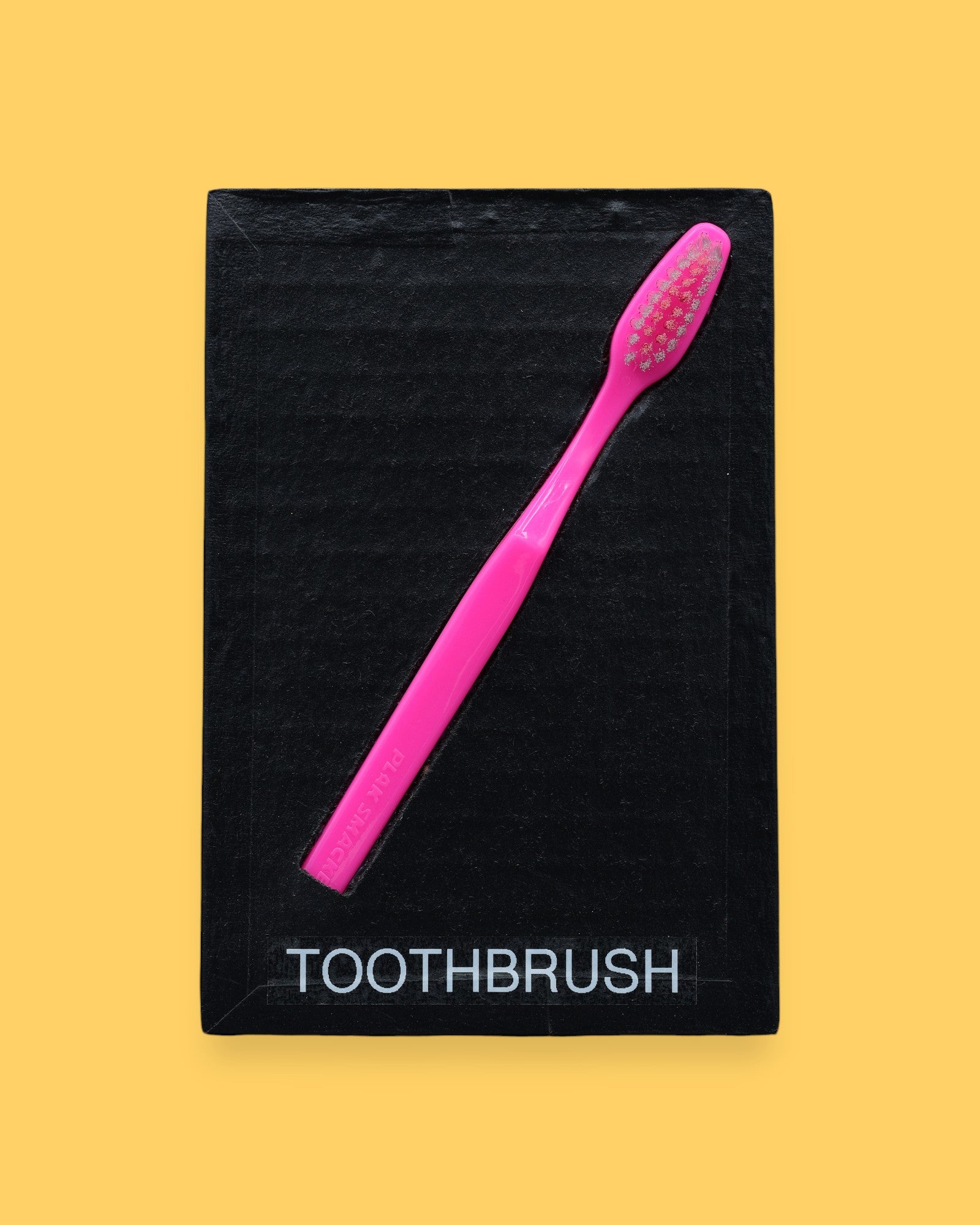 The Toothbrush symbol has a plastic toothbrush is embedded in a diagonal position from the top right corner to the bottom left, with the tactile bristles at the top. Labeled "Toothbrush" in black font on white backing at the bottom of the symbol. The background of the symbol is white. 