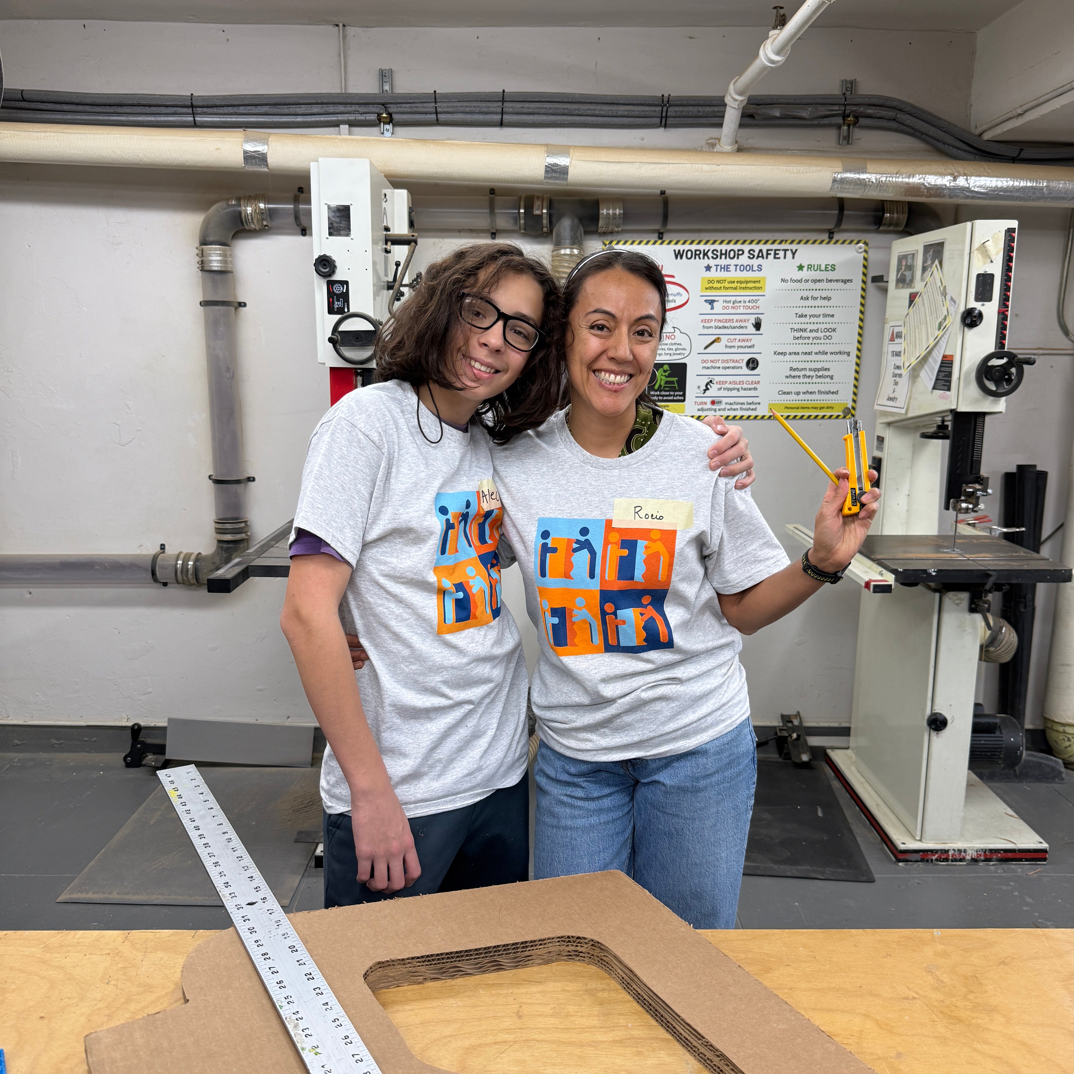 Two people standing together in the Adaptive Design a workshop. They are both wearing ADA t-shirts and jeans, and are posing happily. One person has curly dark hair and glasses, and the other has pulled back hair. They are standing close together and smiling. Around them is a workbench area with tools and equipment. On the right side, there is a large piece of cardboard or wood with a long ruler or measuring stick placed across it, getting ready for a volunteer build workshop. 