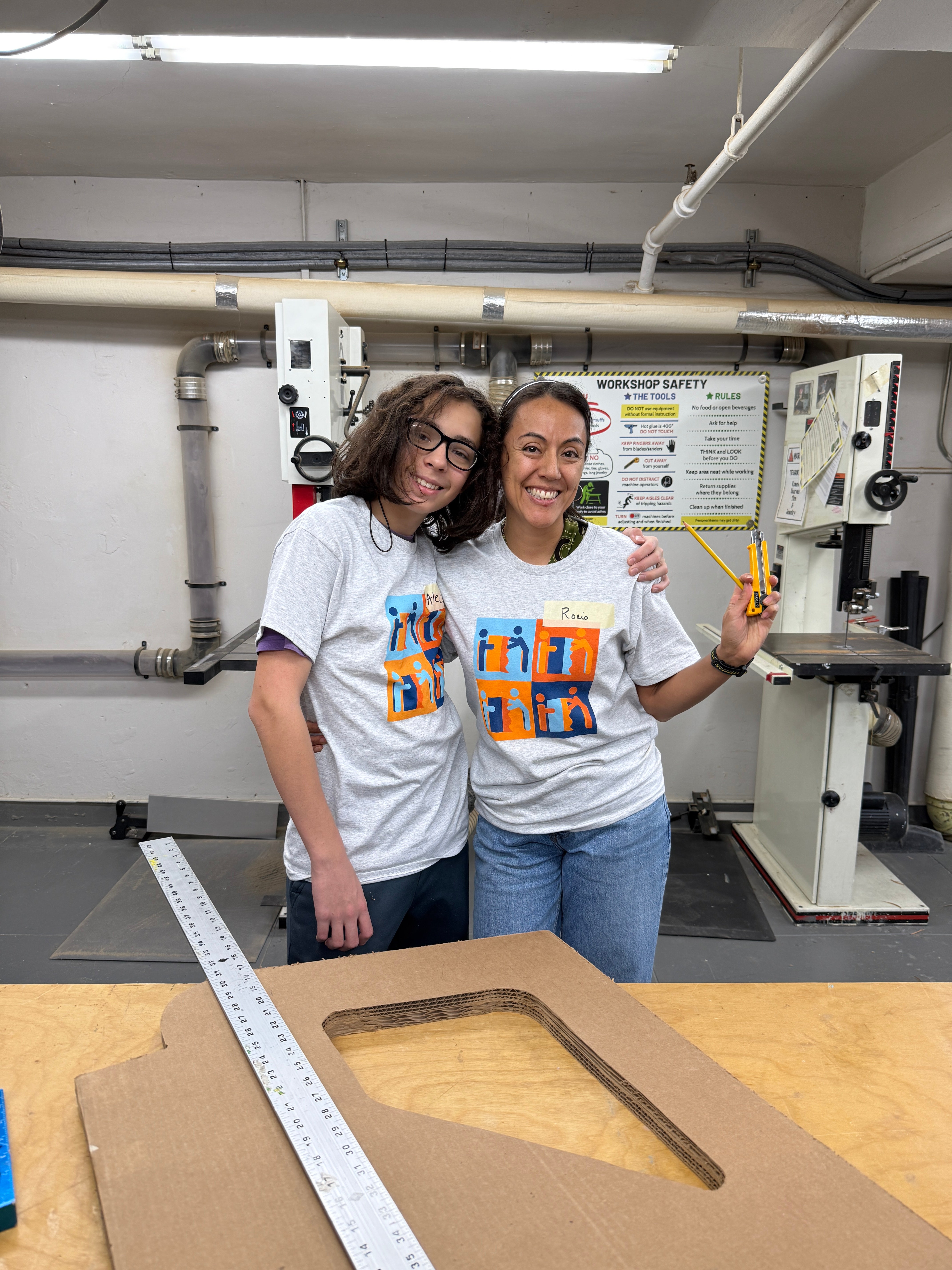 Two people standing together in the Adaptive Design a workshop. They are both wearing ADA t-shirts and jeans, and are posing happily. One person has curly dark hair and glasses, and the other has pulled back hair. They are standing close together and smiling. Around them is a workbench area with tools and equipment. On the right side, there is a large piece of cardboard or wood with a long ruler or measuring stick placed across it, getting ready for a volunteer build workshop. 
