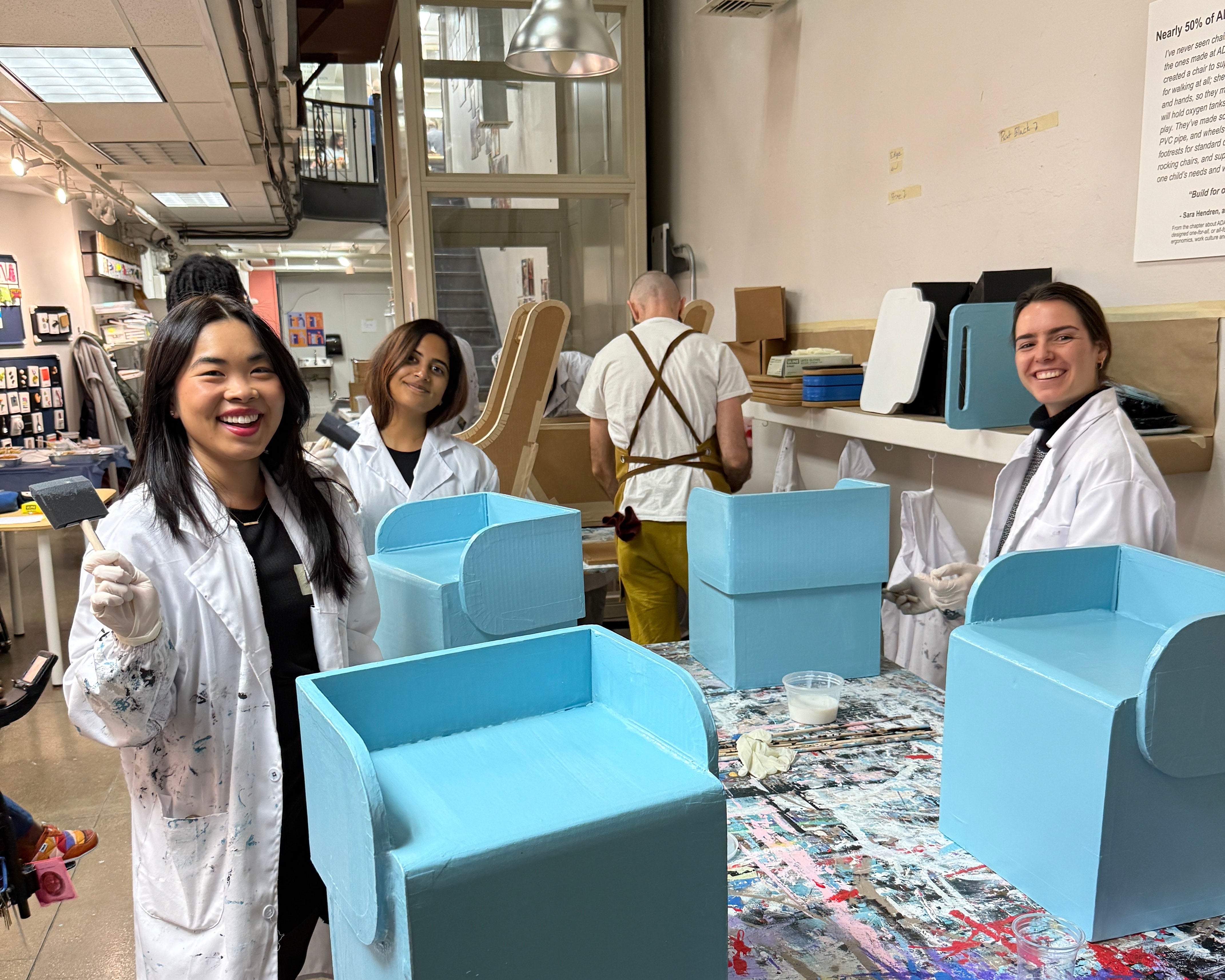 People in a workshop setting are painting blue chairs and a colorful table.