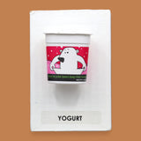 The Yogurt tactile symbol with faux food yogurt container embedded in the center. Labeled "Yogurt" in black font on white backing at the bottom of the symbol. The background of the symbol is white. 