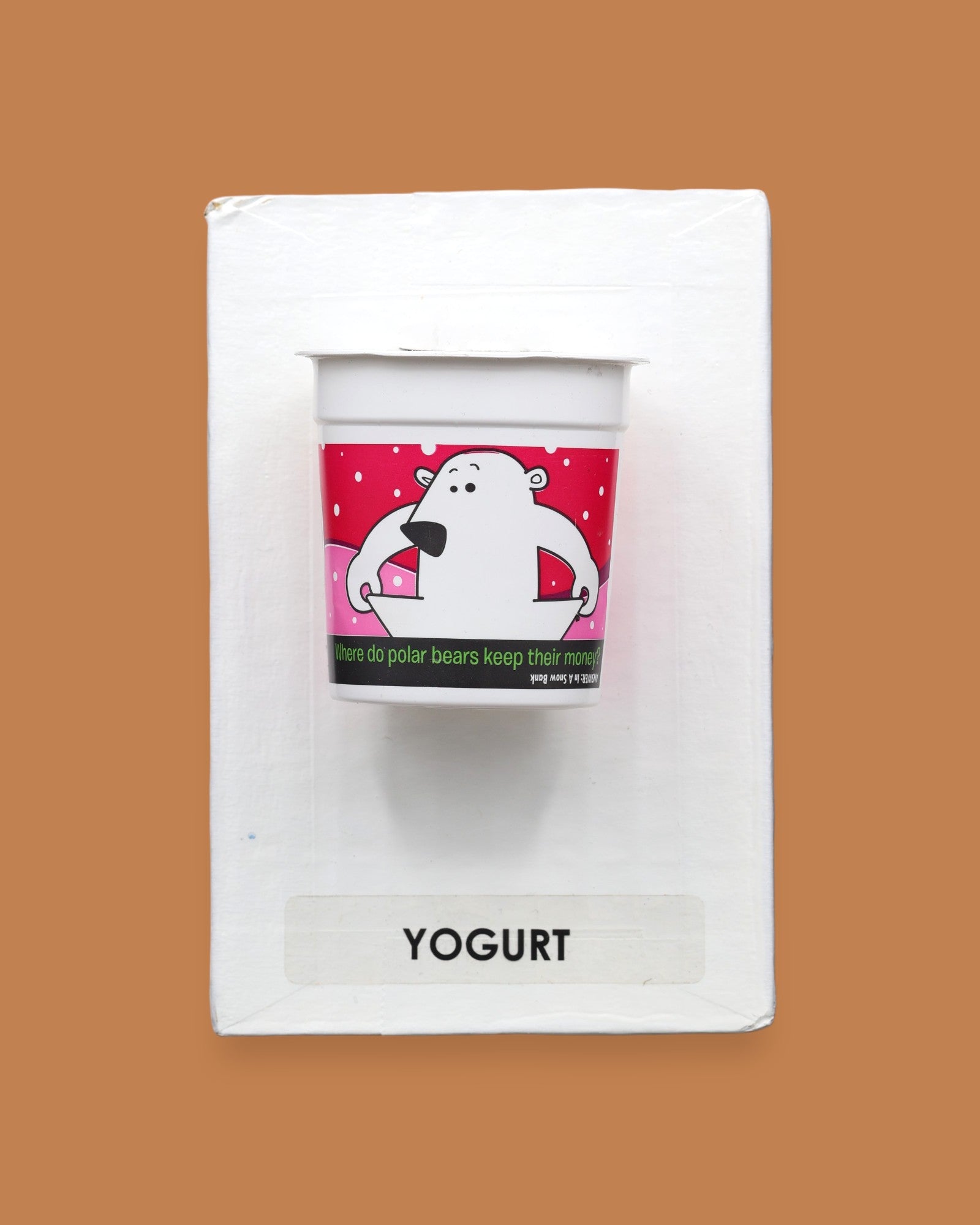 The Yogurt tactile symbol with faux food yogurt container embedded in the center. Labeled "Yogurt" in black font on white backing at the bottom of the symbol. The background of the symbol is white. 